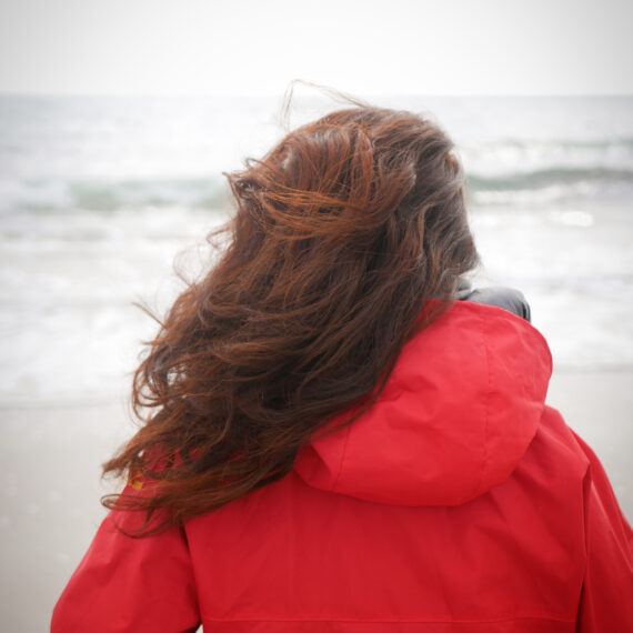 femme de dos en ciré rouge, longue chevelure rousse qui regarde la mer