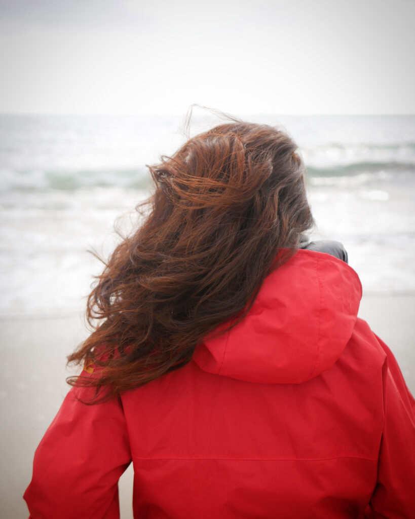 femme de dos en ciré rouge, longue chevelure rousse qui regarde la mer